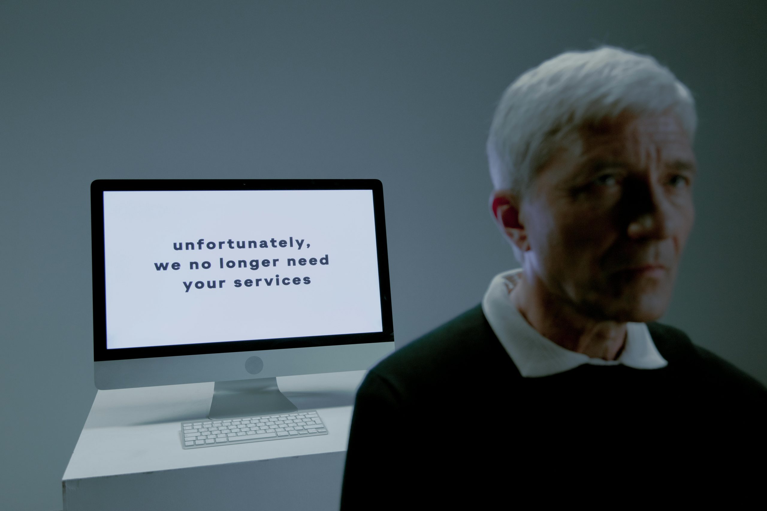 professionally dressed man walking away from a computer monitor displaying 'unfortunately, we no longer need your services'